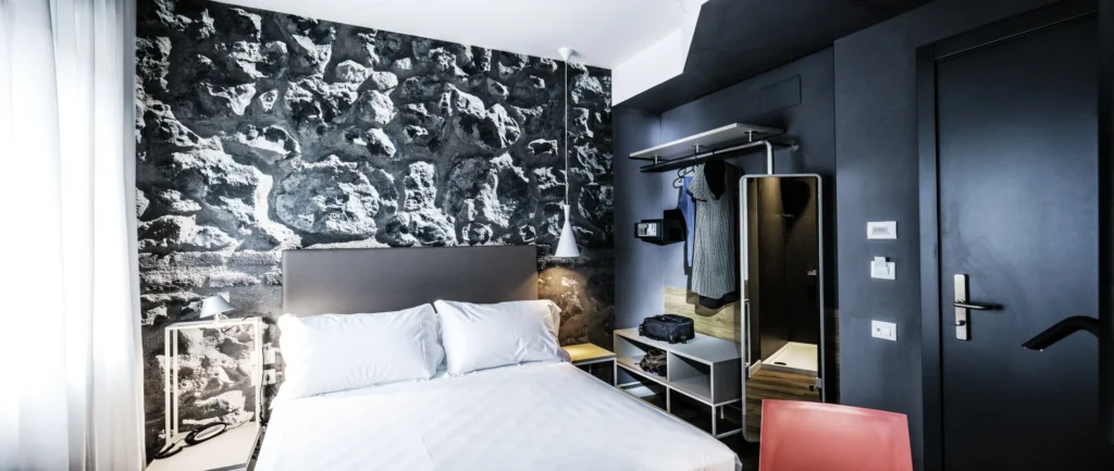 Nell'immagine, la camera Standard dell'Hotel Corsi: si vede il letto matrimoniale e, di fianco, un guardaroba a vista e uno specchio a figura intera. Sulla destra, la porta della camera.