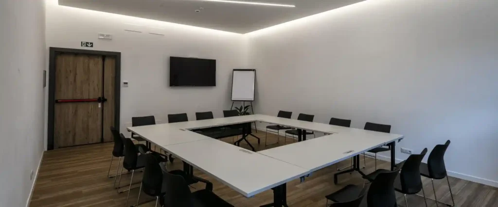 Nell'immagine, la sala meeting dell'Hotel Corsi: si vede un tavolo circondato da sedie e una TV appesa alla parete. Di fianco alla TV, una lavagna a fogli mobili.